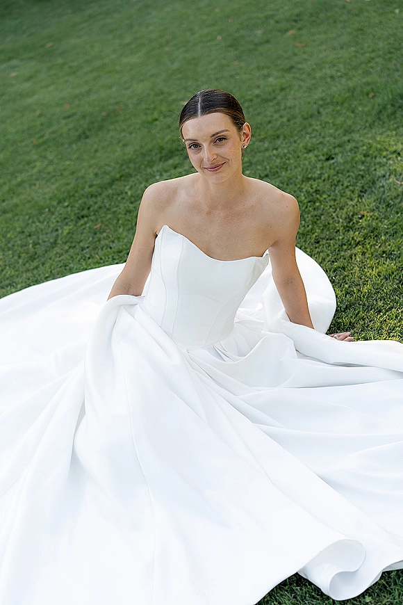 Bridal portrait of a bride in a strapless wedding dress with full skirt and train, seated on a green grass lawn, stud earrings visible
