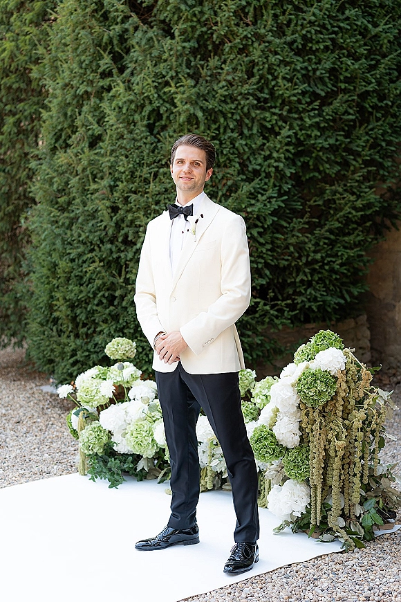 Groom portrait in a white tuxedo jacket with black bow tie and boutonniere, hands clasped beside aisle florals and evergreen hedge