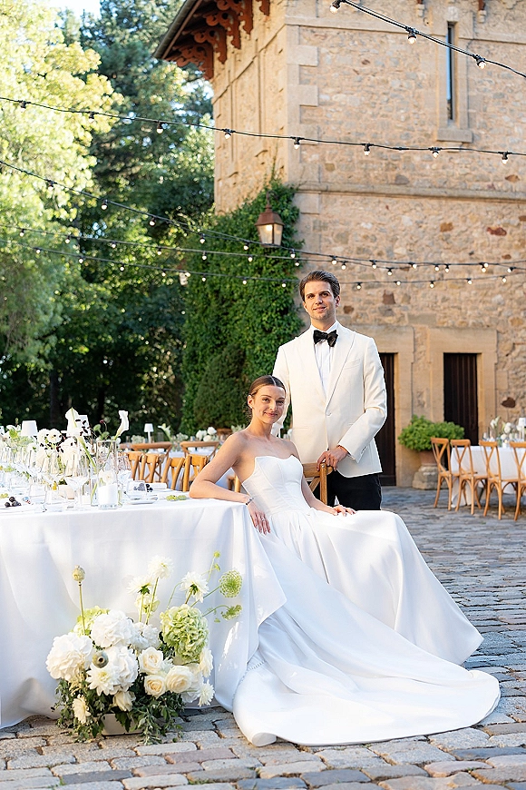 Couple portrait of bride seated in a strapless wedding dress with long train and groom in white tuxedo at candlelit courtyard table under string lights