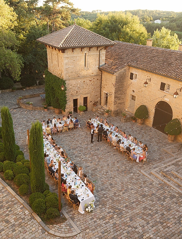 Outdoor wedding reception in a stone courtyard with long banquet tables, cross-back chairs, string lights, and topiary in terracotta pots