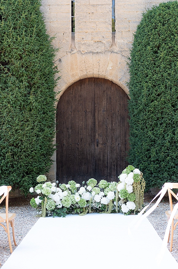 Ceremony altar decor with a ground floral altar of white hydrangeas beside a white aisle runner, set before a stone archway doorway.