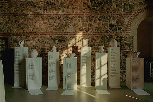 Wedding seating chart scrolls with calligraphy lettering on white plinths, styled with ceramic vases against a stone brick wall and arched doorway