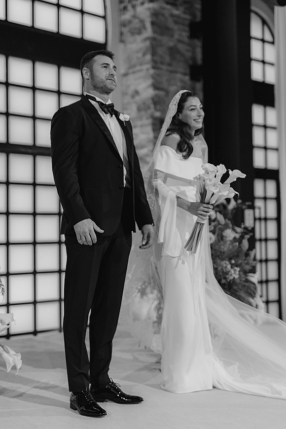 Ceremony moment as bride in off the shoulder wedding dress holds calla lilies beside groom in tuxedo, veil accenting vows by grid windows
