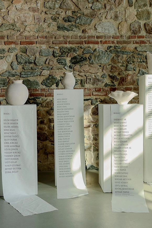 Wedding seating chart modern seating chart display on white pedestal stands with printed guest list cards and ceramic vases against a sunlit stone wall