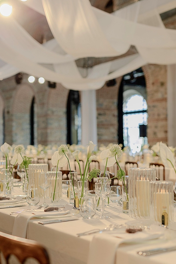 Reception tablescape with white wedding tablescape details, bud vases of calla lilies and ribbed glass candles in a brick, arched-window venue