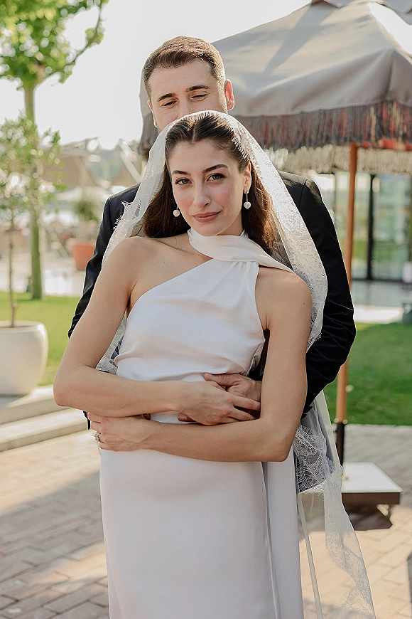 Couple portrait of bride and groom embrace, groom hugging bride as she looks at camera, bridal veil flowing on a patio with greenery