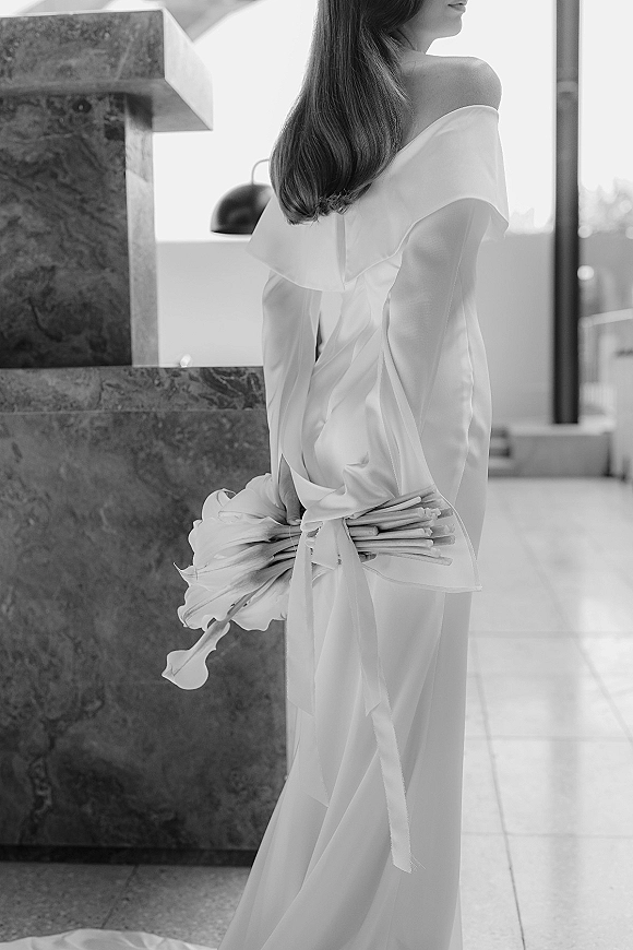 Bridal portrait of a bride in an off the shoulder wedding dress holding a calla lily bouquet with ribbon in a window-lit stone venue