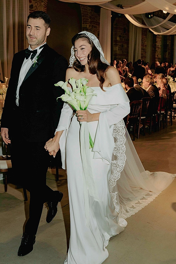 Wedding recessional as bride and groom walking hand in hand, bride holding calla lilies with veil trailing in a draped-ceiling reception hall
