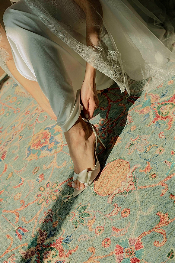 Bridal shoes, ivory slingback wedding heels with bow detail under lace-trim veil on a patterned vintage rug with sunlight shadows