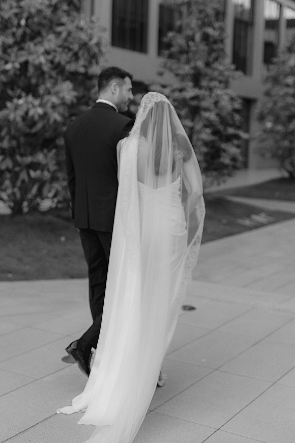 Couple portrait of bride and groom walking away on a city sidewalk, her lace wedding veil and dress train flowing behind him in tuxedo