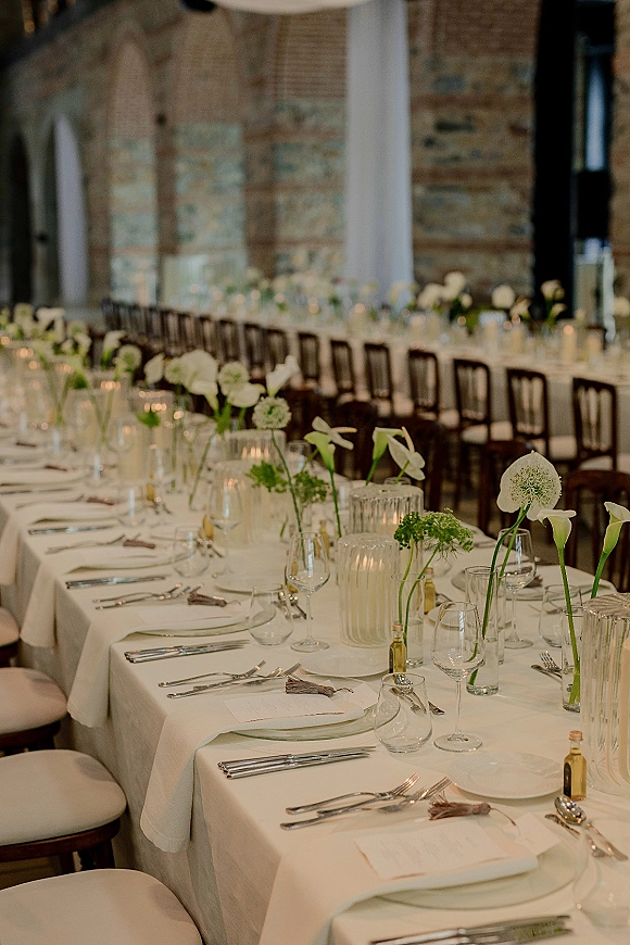 Reception tablescape with long banquet table wedding details, white tablecloth, clear glass plates, calla lilies and votive candles by brick wall