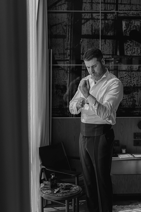 Groom getting ready, adjusting cufflinks in a white dress shirt by a window, cologne on a side table in soft natural light