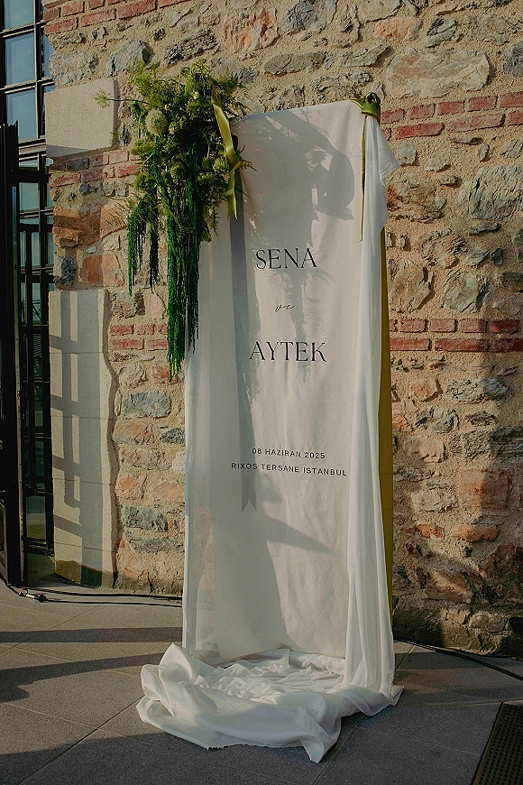 Wedding welcome sign with white draped fabric and greenery arrangement, green ribbon tied, set by a sunlit stone wall entrance
