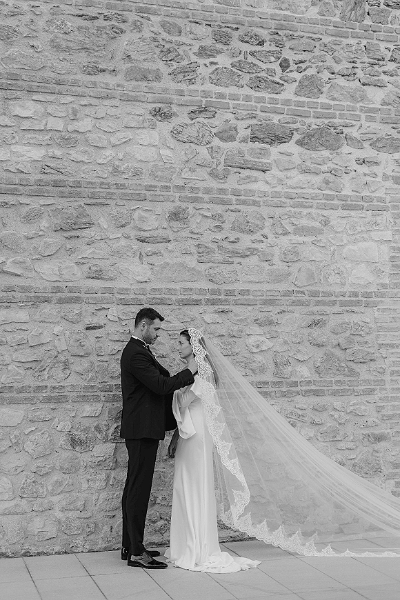 Couple portrait in a black and white wedding portrait, bride and groom touching foreheads by a stone and brick wall, long lace veil flowing