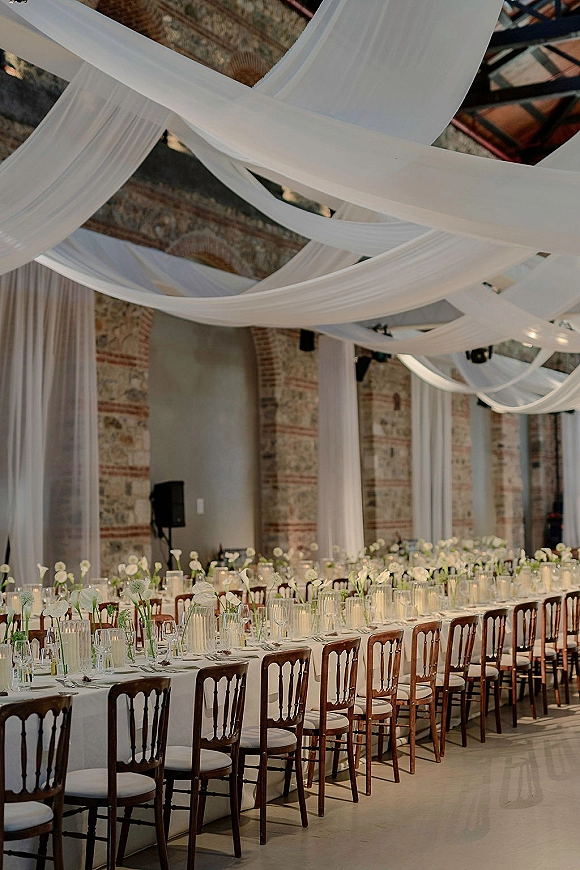 Reception tablescape with a long banquet table wedding setup, white linens, glassware, candles, and tall white florals beneath draped ceiling in brick hall