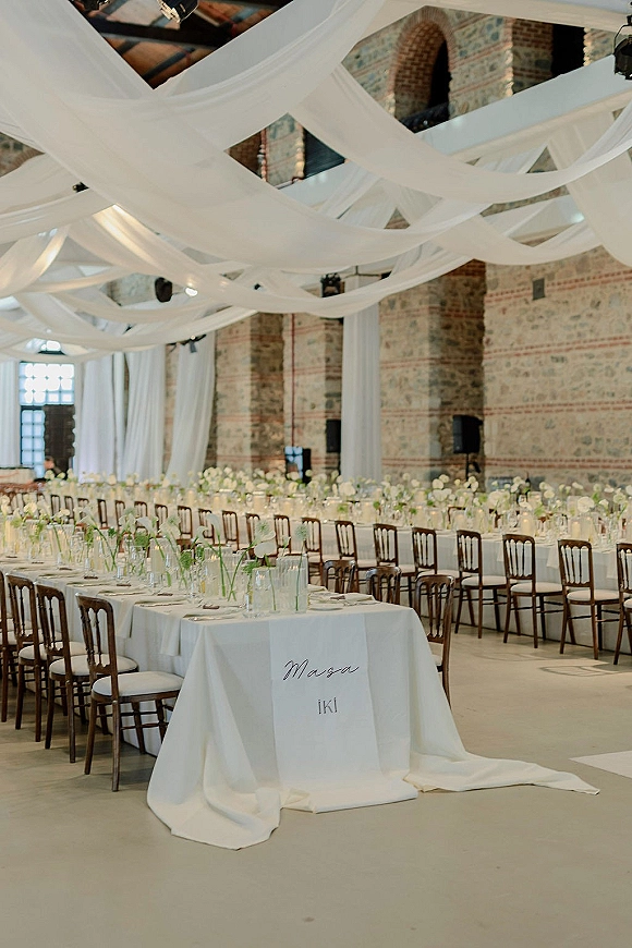 Reception tablescape with white draped ceiling decor, long banquet tables, and green-and-white florals in a brick hall with arched openings