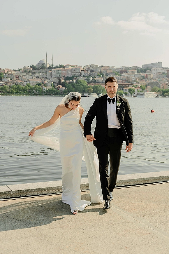 Couple portrait of bride and groom walking hand in hand, her long veil flowing, with waterfront city skyline and mosque domes behind