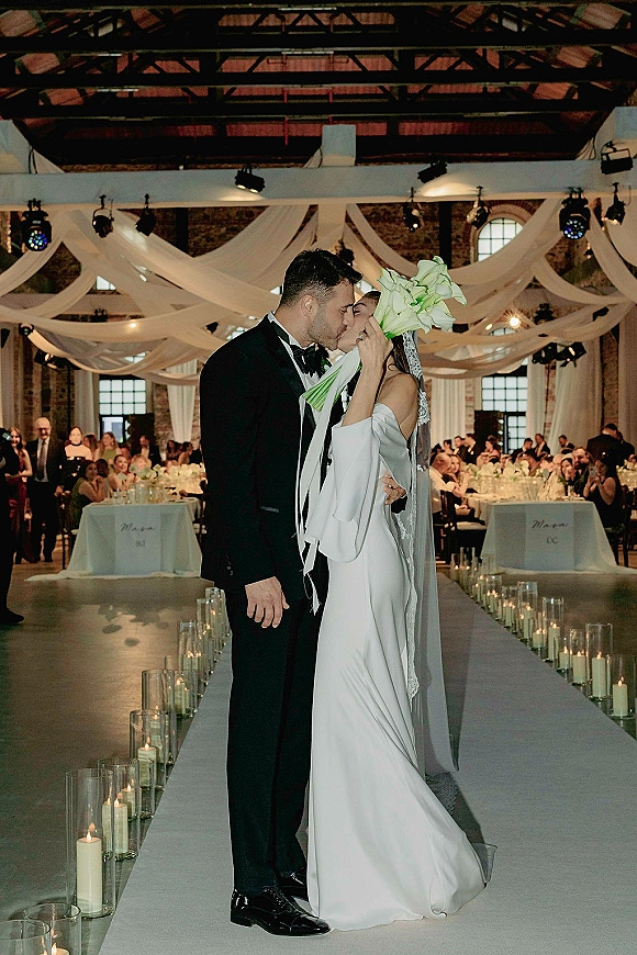 Wedding kiss portrait of bride and groom kiss, her long sleeve gown and veil with white bouquet, under bistro lights in a brick-beam hall