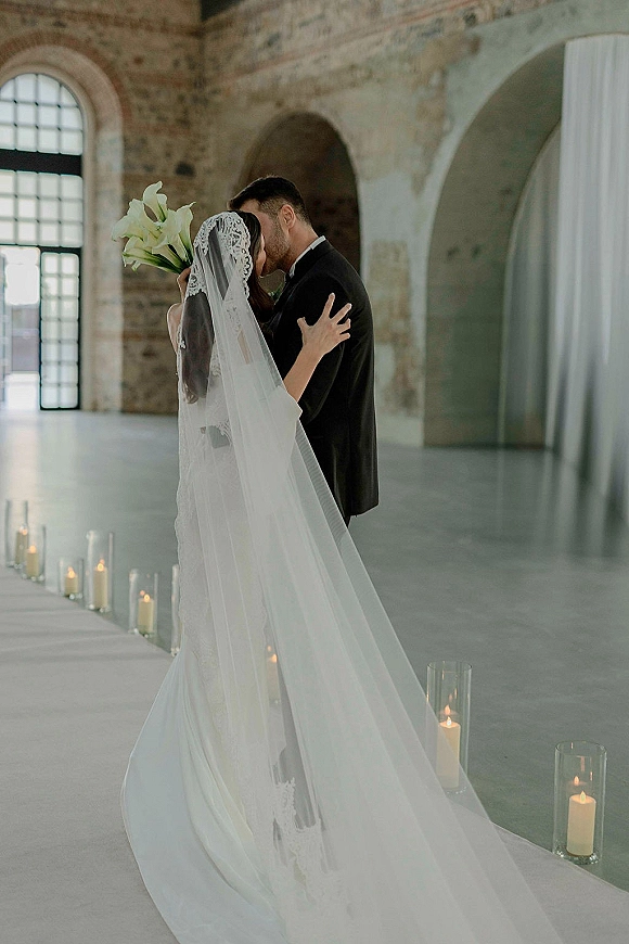 Wedding kiss portrait of bride and groom kissing, bride holding calla lilies, lace veil flowing in an industrial loft with brick wall and candles