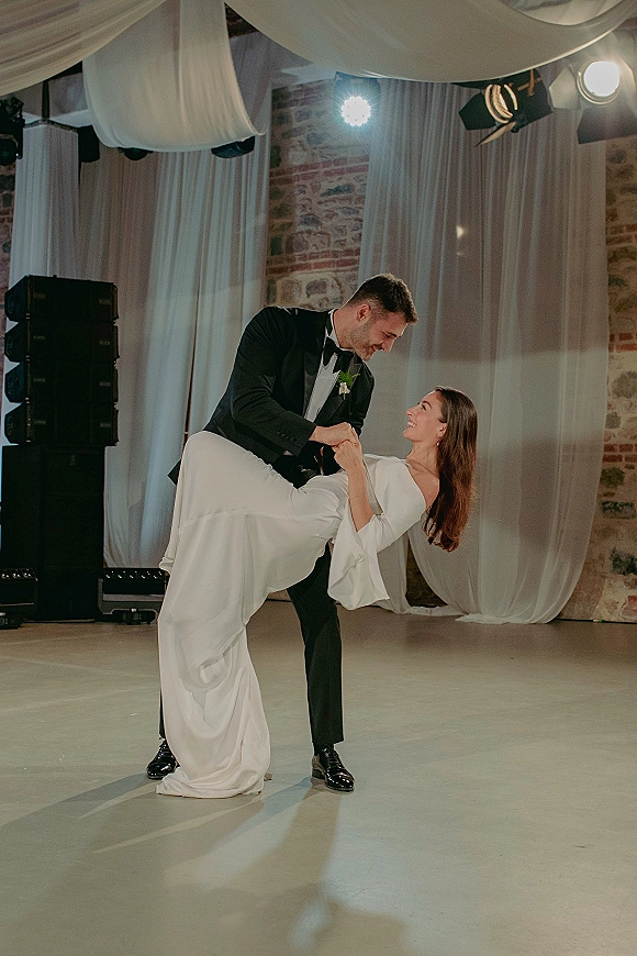 First dance moment as bride in a long sleeve wedding dress dips with groom in black tuxedo under draped ceiling, brick wall behind