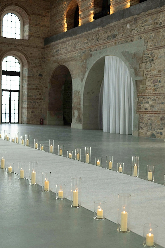 Ceremony aisle decor with a white aisle runner and pillar candles in glass cylinders, set in a stone hall with arched windows