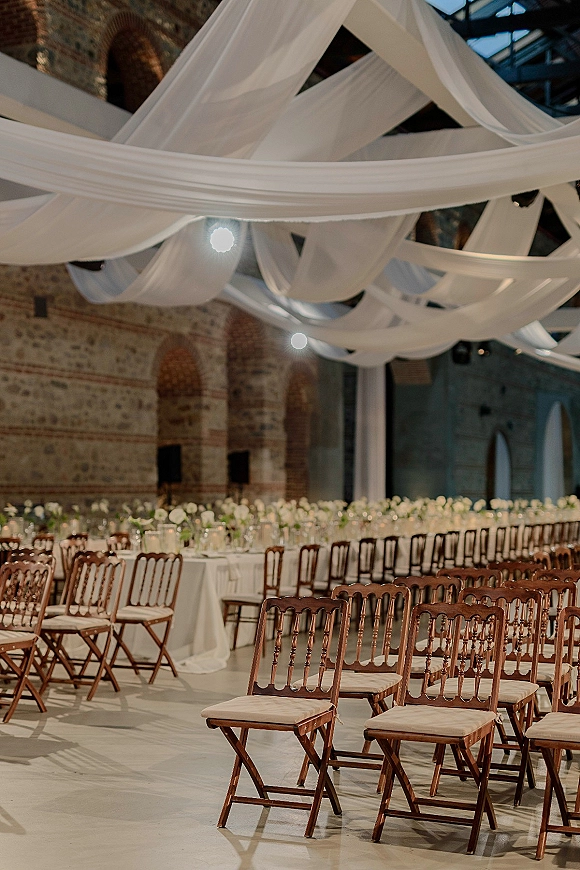 Reception tablescape with white draped ceiling decor over round tables, low floral centerpieces, and votive candles in an industrial brick hall