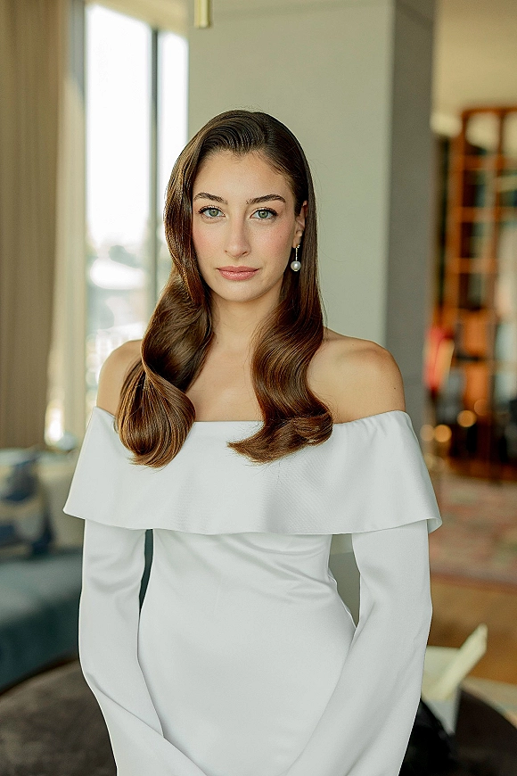 Bridal portrait of a bride in an off the shoulder wedding dress with pearl drop earrings, posed in soft window light by curtains and a bookshelf