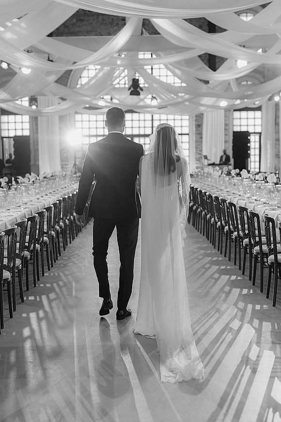Wedding recessional with newlyweds walking away between banquet tables, bride’s long veil trailing in sunlit brick-walled hall