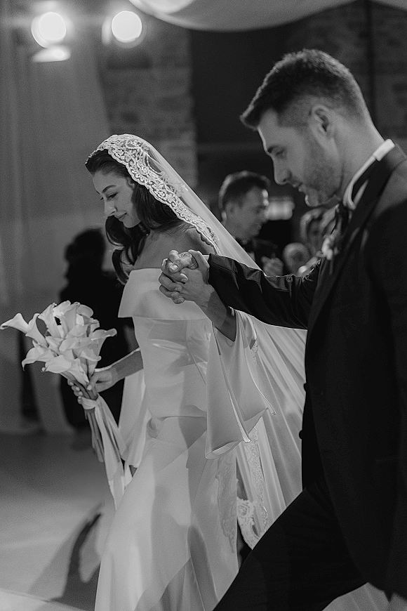 Ceremony moment as bride and groom holding hands, her lace mantilla veil and calla lily bouquet in a softly lit indoor aisle with guests