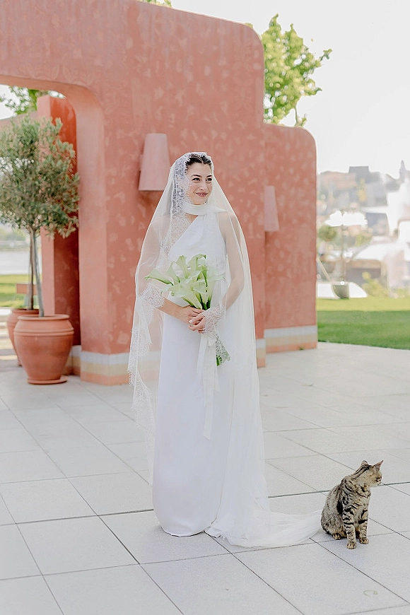 Bridal portrait of a bride holding bouquet with lace veil and calla lilies, standing by a terracotta stucco wall on a tiled patio