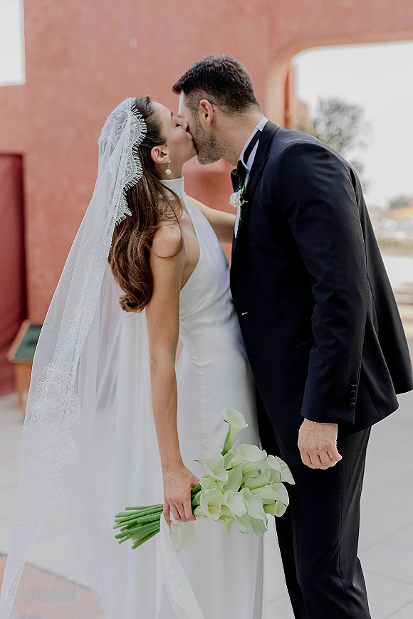 Wedding kiss portrait of bride and groom kiss as she holds a calla lily bouquet, veil flowing by a stucco wall outdoor walkway