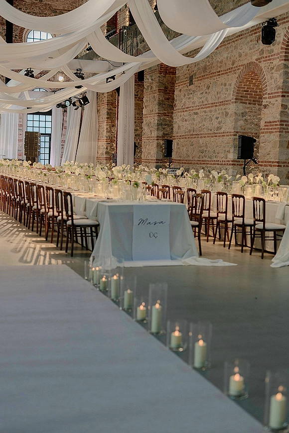 Reception tablescape with long banquet table wedding setup, white floral centerpieces, glassware and votive candles under draped ceiling in brick loft venue