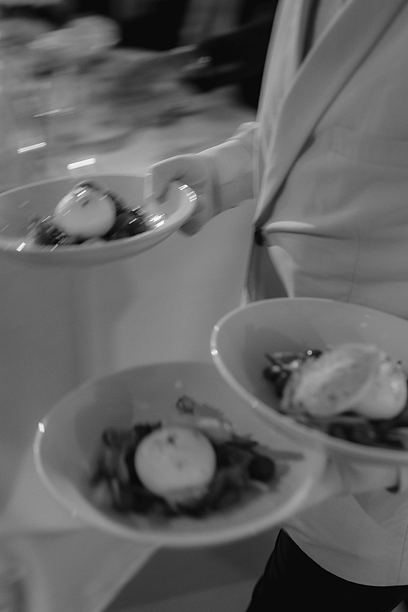 Wedding dinner service as a server carries white bowls of plated salad with leafy greens past glassware at an indoor reception table