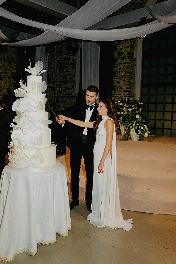 Wedding cake cutting with couple cutting wedding cake beside a tall tiered cake, groom in tuxedo and bride in gown under draped ceiling