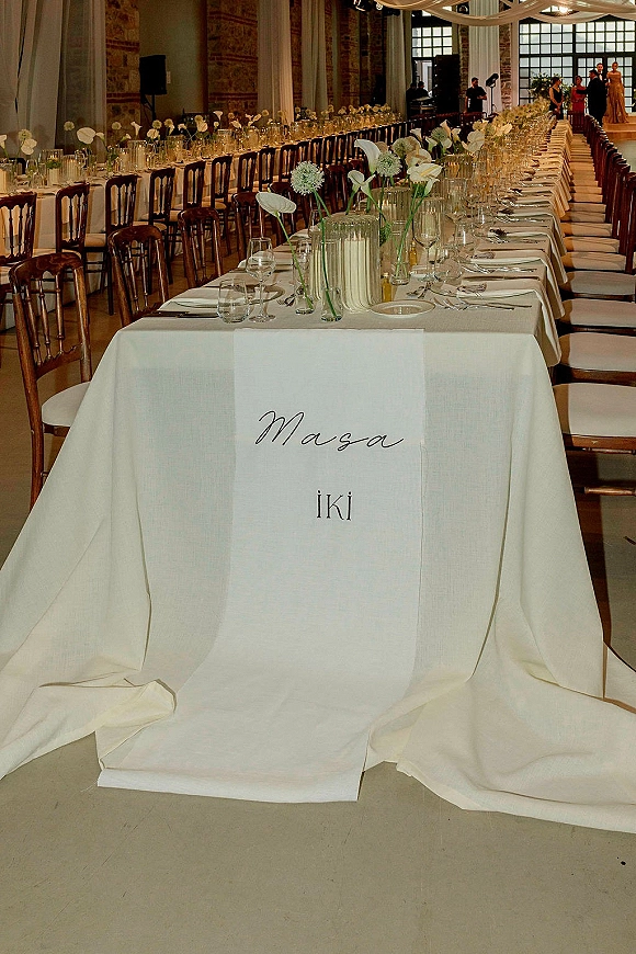 Wedding reception tablescape with a long banquet table wedding setup, white linens, calla lilies in glass vases, and draped ceiling in a stone hall