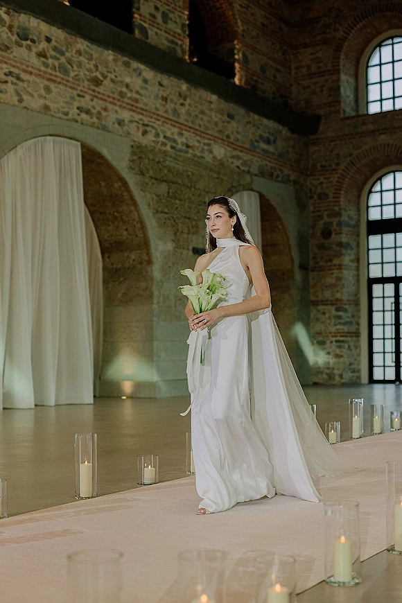 Bridal portrait of a bride holding bouquet of white calla lilies in a veil, walking an aisle runner with glass candles in an arched stone hall