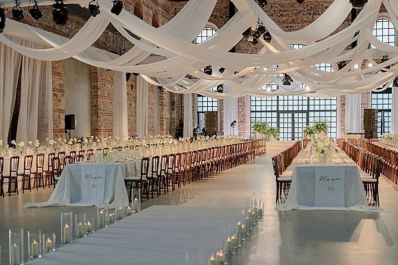 Reception tablescape with long banquet table wedding details, greenery centerpieces and taper candles beneath a white draped ceiling in brick loft hall