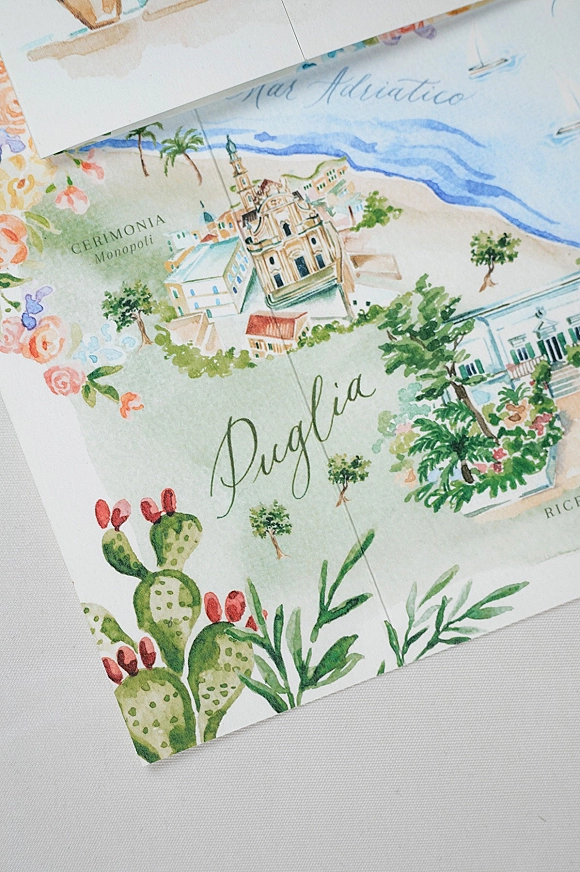 Wedding invitation with watercolor wedding invitation floral border, map artwork and calligraphy lettering beside an envelope on light fabric surface