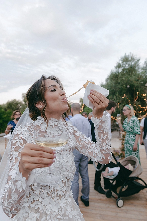 Bride candid moment as she sips champagne and takes an appetizer bite, lace long-sleeve dress and veil under string lights outdoors
