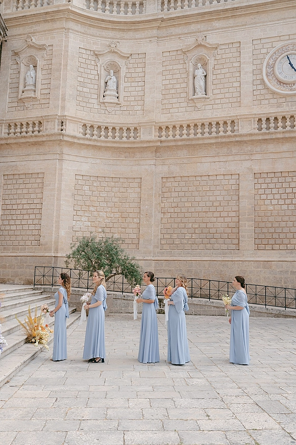 Bridesmaid processional with bridesmaids walking down aisle in light blue dresses, holding bouquets in a stone courtyard by a grand facade