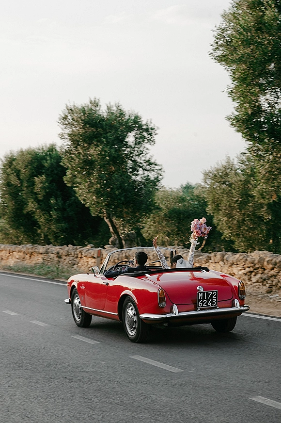 Wedding getaway car just married car with bride in veil holding bouquet in a red vintage convertible on a country road by stone wall