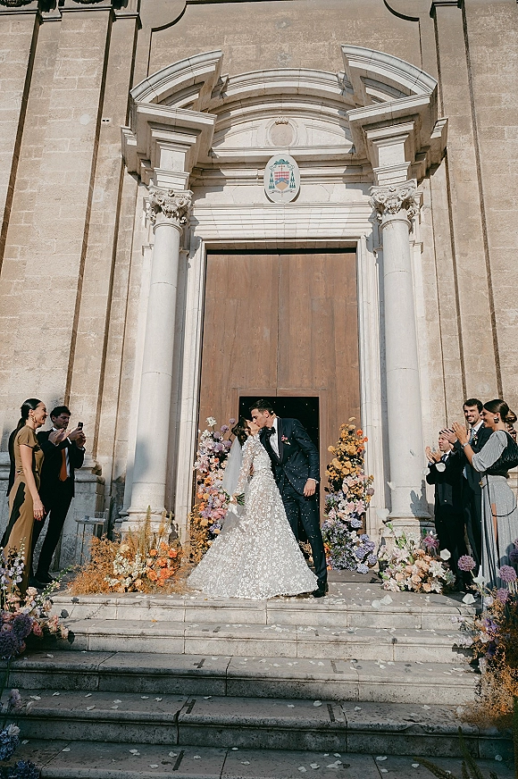 Wedding kiss as the bride in a lace dress and veil and groom in a tuxedo share a just married kiss on stone church steps amid petals