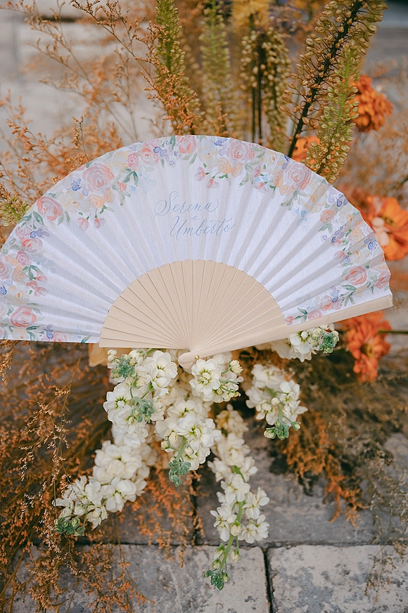 Wedding fan with personalized calligraphy and floral pattern, wooden ribs and white flowers with dried foliage on stone pavement