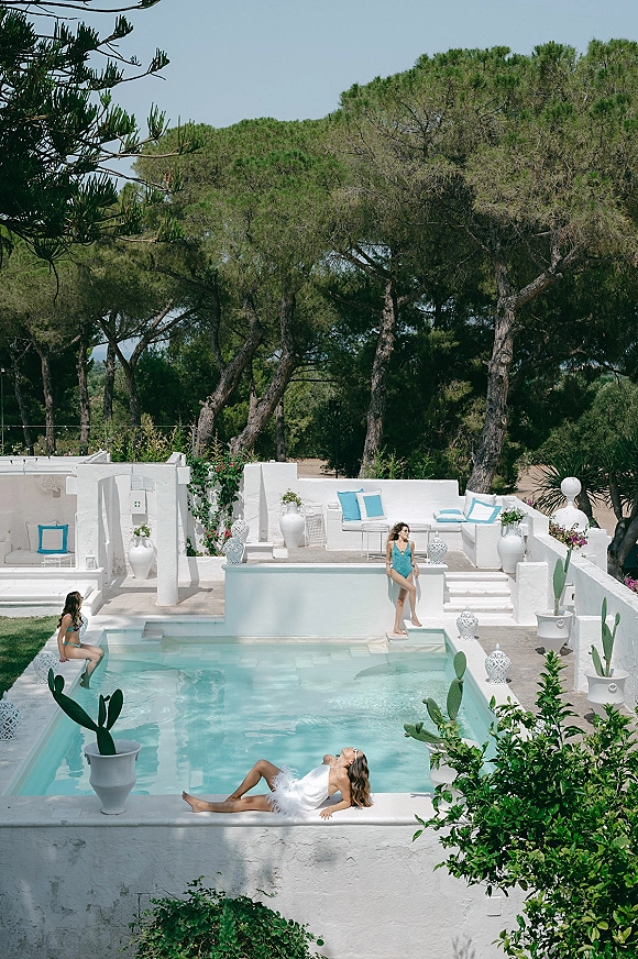 Poolside bridal portrait of a bride by the pool in a feather-trim bridal mini dress, lounging on a white stucco terrace by turquoise water
