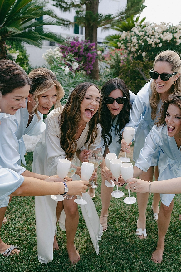 Bridesmaids toast with champagne flutes during a getting ready toast, wearing satin robes and sunglasses on a garden lawn with palms