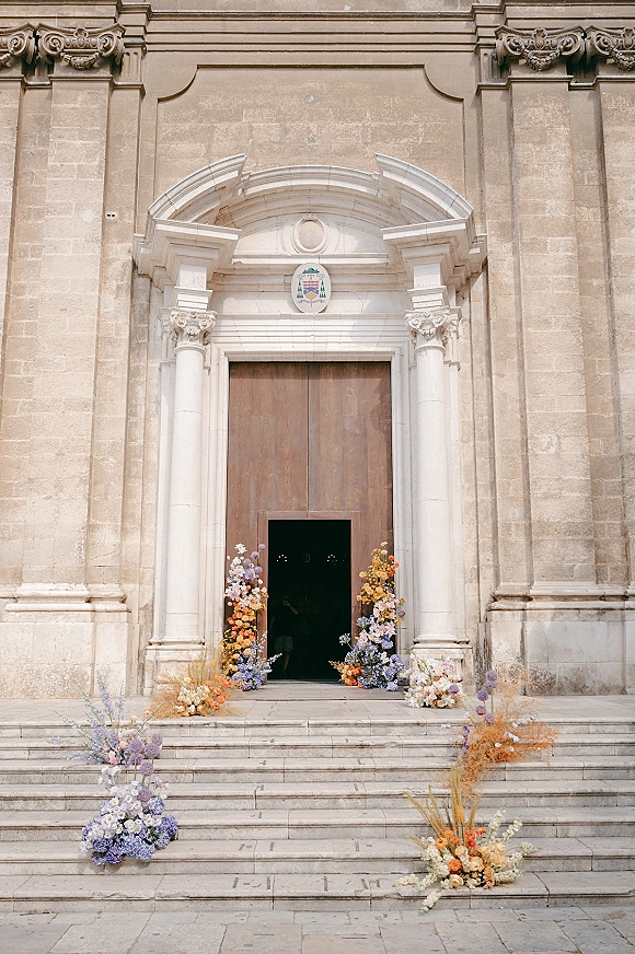 Ceremony entrance decor with church entrance flowers framing a stone arched doorway, pastel lavender and orange florals and dried grasses on steps