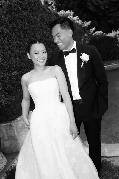 Couple portrait of bride and groom smiling, her strapless lace ballgown, his classic look with a black bow tie, in a garden by a stone wall.