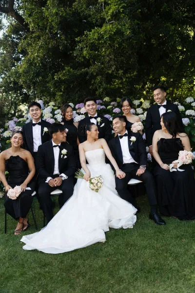 Wedding party portrait with the bride and groom seated, friends gathered behind, hydrangea bushes in a garden, bow ties adding classic polish.