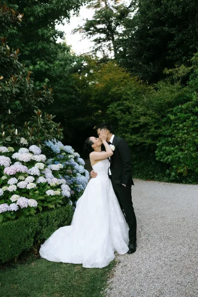 Wedding kiss portrait as a bride and groom embrace beside blooming hydrangeas on a garden path beneath trees and hedges, tender and intimate.