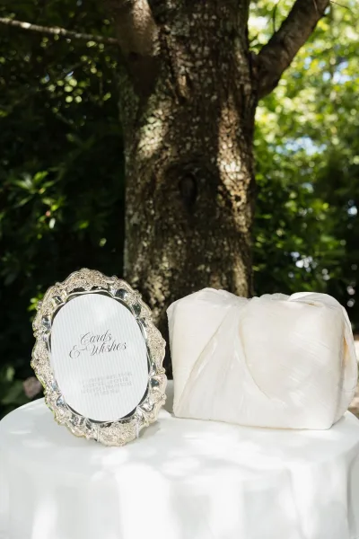 Wedding card table with gift table signage in a silver frame, positioned beside a tree trunk amid garden greenery.